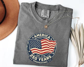 May include: A gray Comfort Colors t-shirt with a round graphic featuring the American flag and the text "AMERICA 250 YEARS SEMIQUINCENTENNIAL EST. 1776". A silver necklace with a pendant is on the shirt. A brown hat is visible.
