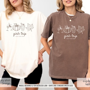May include: Two oversized t-shirts, one ivory and one espresso, each featuring a white graphic of wine glasses, a pumpkin, a beverage, and a leaf. The text "girls trip NEW YORK 2000" is printed below the graphic. The shirts are unisex sizes.
