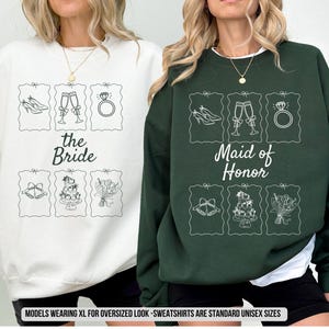 May include: White and forest green sweatshirts with bridal-themed illustrations. The white sweatshirt says "the Bride" and the green one says "Maid of Honor". Each shirt features drawings of wedding elements.