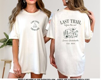 May include: Two white t-shirts with a camping theme. The front shirt says "Camp Emma Grand Lake" with a campfire graphic. The back shirt says "Last Trail Before the Veil Emma's Bachelorette Est. 2024" with a mountain and tent graphic.
