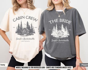May include: Ivory and pepper-colored t-shirts with the text "CABIN CREW" and "THE BRIDE" printed on them. Each shirt features a cabin graphic with trees and the text "Sarah's Bachelorette Lake Panache". The shirts are standard unisex sizes.