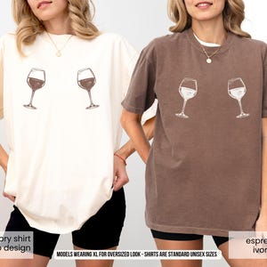 Könnte beinhalten: Zwei übergroße T-Shirts, eines in Elfenbein und eines in Espresso, jeweils mit einem Weinglas-Design. Das elfenbeinfarbene Shirt hat ein dunkelweinrotes Design, während das Espresso-Shirt ein weißes Design hat. Die Shirts sind in Standard-Unisex-Größen erhältlich.