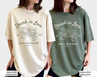 May include: Ivory and moss green t-shirts featuring the text "Drunk in Love" and "Emma's Bachelorette Newport, RI". The design includes two champagne glasses with bows and the year 2026.