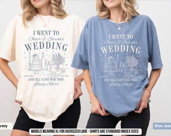 May include: Ivory and blue jean t-shirts with the text "I Went to Oliver & Sarah's Wedding" and "And All I Got Was This Lousy Shirt". The shirts have a graphic of a wedding cake, wine glasses, and flowers.