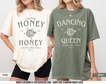 May include: Two t-shirts, one ivory and one moss green, with a disco ball graphic and text that reads "I Found My Honey" and "Dancing Queen". The text also includes "Sarah's Last Disco" and "Miami".