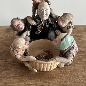 May include: A ceramic figurine of a man with five monkeys surrounding him. The man is wearing a black and white robe with a floral pattern. The monkeys are all different colors and have different expressions. The figurine is sitting on a woven basket.