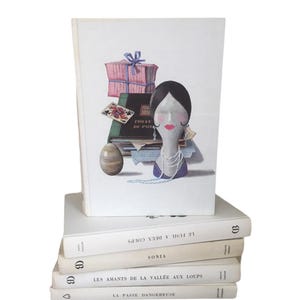May include: A stack of five vintage paperback books with white covers and black text. The top book features a colorful illustration of a woman with a pink box, a deck of cards, and a book.