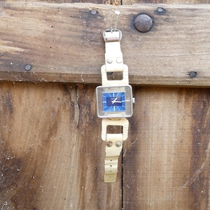 Könnte beinhalten: Eine weiße Lederarmbanduhr mit einem quadratischen blauen Zifferblatt und silbernen Zeigern. Die Uhr hängt an einer Holzoberfläche.