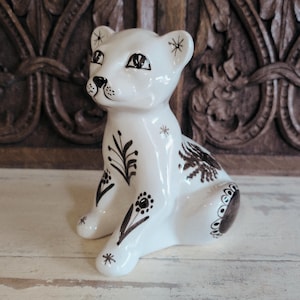 May include: A white ceramic figurine of a seated big cat, featuring hand-painted dark brown floral and star motifs. The feline has pointed ears, almond-shaped eyes, and a smooth, glossy finish, with detailed botanical patterns on its legs and back.