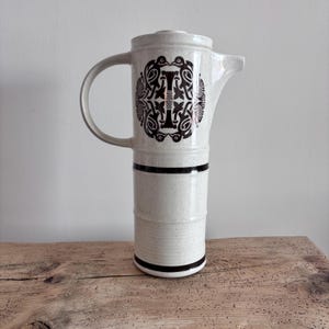 May include: A tall, cream-colored ceramic coffee pot with a black decorative pattern and a handle. The pot has a cylindrical body with black bands and a lid. The pot is sitting on a wooden surface.