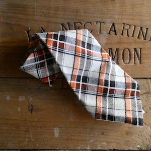 Może przedstawiać: Krawat w kratę w kolorze białym, pomarańczowym i czarnym. Krawat jest złożony i leży na drewnianej powierzchni.