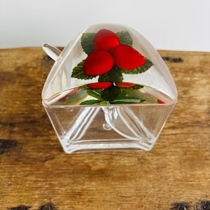 Könnte beinhalten: Eine transparente, geometrisch geformte Zuckerdose mit Deckel und Löffel. Der Deckel zeigt drei rote Erdbeeren mit grünen Blättern, die in transparentem Material eingeschlossen sind. Die Schale steht auf einer Holzoberfläche.