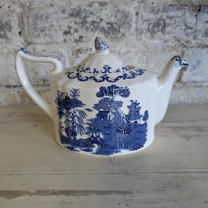 Può includere: Teiera in ceramica bianca con un motivo blu Willow. La teiera presenta un manico curvo, un beccuccio con un dettaglio a forma di uccello e un coperchio con un pomello decorativo. Il design blu e bianco raffigura una scena paesaggistica.
