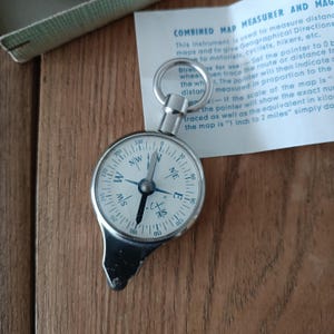 May include: A silver compass with a white face and black markings. The compass has a magnifying glass on the side and a ring at the top. The compass is labelled "Combined Map Measurer and Magnifier" and "C. & S.  3/S".