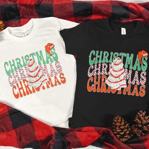 May include: Two short-sleeved t-shirts, one white and one black, with a Christmas-themed graphic. The design features the word "CHRISTMAS" in green, red, and orange, along with a decorated Christmas tree and a Santa hat.