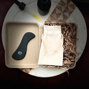 Puede incluir: Una herramienta de gua sha negra en una caja de cartón, una pequeña bolsa blanca con cordón y una botella de vidrio con gotero.