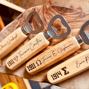 May include: Four wooden bottle openers with stainless steel tops are displayed on a wooden cutting board. Each opener has a different name and year engraved on the wooden handle. The names are: Shaun Wimbash, Terence Carter, James E. Clyburn, and Eric Ross.