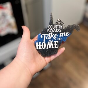 May include: A West Virginia state-shaped car air freshener. The air freshener is black and blue with the words "COUNTRY ROADS Take me HOME" in white lettering. It has a silver metal ring for hanging.