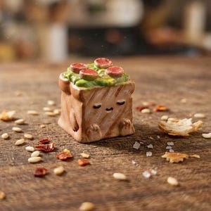 May include: A small, square-shaped ceramic figurine resembling a piece of toasted bread with grill marks. The top is covered in green textured topping and four small red circular slices. It features a simple hand-painted face with closed eyes.