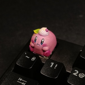 May include: A pink, cartoon-style keycap featuring a character with large blue eyes, small arms, and feet. The keycap has a whipped cream-like topping with a green leaf. It is placed on a black keyboard, near the number keys.