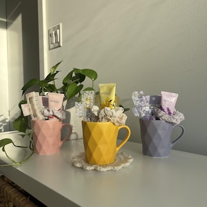 May include: Three decorative mugs filled with beauty products. A pink, yellow, and purple mug, each with a geometric design. The mugs contain items such as hair ties and skincare products. The yellow mug sits on a crocheted coaster.