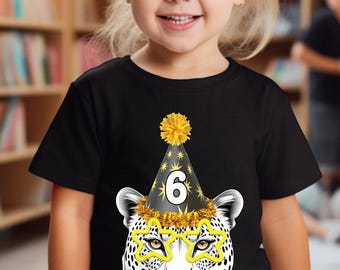 May include: A black t-shirt featuring a graphic of a leopard wearing a party hat with the number 6 and star-shaped glasses. The leopard's face is white with black spots. A yellow pom-pom is on top of the hat. The shirt is worn by a young girl.