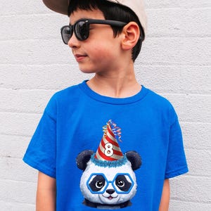 May include: A young person wearing a blue t-shirt featuring a panda graphic. The panda is wearing glasses and a party hat with the number 8. The person is also wearing sunglasses and a baseball cap.
