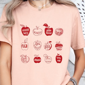 May include: A pink t-shirt with a graphic of red apples and their names, including Granny Smith, McIntosh, Golden Delicious, Cortland, Fuji, Red Delicious, Gala, Braeburn, Pink Lady, Honeycrisp, and  Macoun. The graphic also includes the text "Yummy Time".
