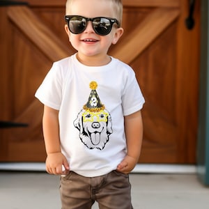 May include: A young boy wearing a white t-shirt with a black and gold dog wearing a party hat and sunglasses. The dog is wearing a party hat with the number "2" on it. The boy is wearing sunglasses and has a spiky hairstyle.