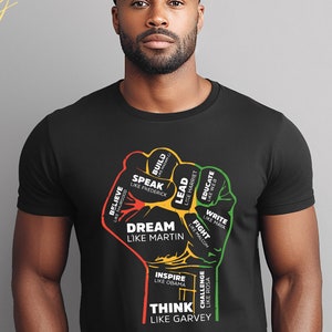 May include: Black t-shirt featuring a colorful fist graphic with the words "Dream Like Martin" and other inspirational phrases. The design includes red, yellow, and green accents, and the text "Think Like Garvey".