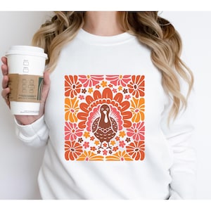 May include: White sweatshirt with a graphic of a brown turkey surrounded by orange, pink, and red flowers. The flowers are arranged in a square pattern.