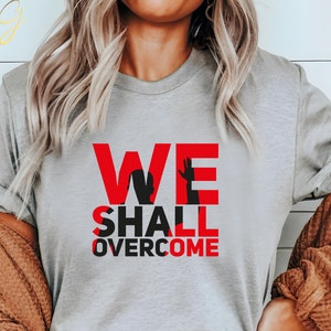 May include: A gray t-shirt with the words "WE SHALL OVERCOME" printed in red. The words are in a bold, blocky font and are centered on the shirt.