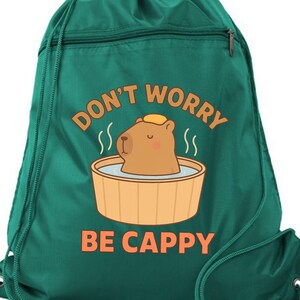 May include: A dark green drawstring backpack featuring a graphic of a brown capybara sitting in a wooden tub with steam rising. The text "DON'T WORRY BE CAPPY" is printed in orange above and below the illustration. The bag has matching green cords.