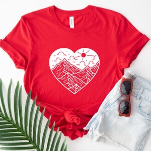 May include: Red t-shirt with a white graphic of a mountain range inside a heart shape. The graphic also includes a sun and clouds.