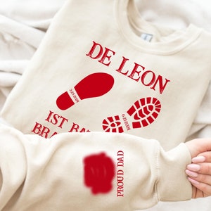 May include: A beige sweatshirt with the text "DE LEON IST RA BR" and two red footprints. The text "PROUD DAD" is on the sleeve.