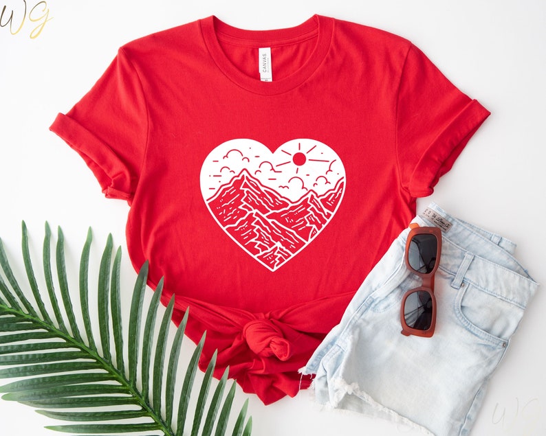 May include: Red t-shirt with a white graphic of a mountain range inside a heart shape. The graphic also includes a sun and clouds.