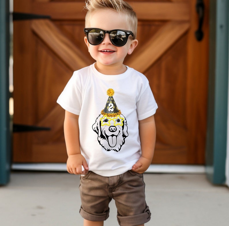May include: A young boy wearing a white t-shirt with a black and gold dog wearing a party hat and sunglasses. The dog is wearing a party hat with the number "2" on it. The boy is wearing sunglasses and has a spiky hairstyle.