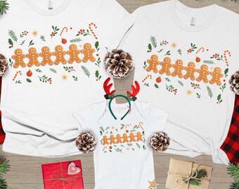 May include: White t-shirts and a onesie with gingerbread man designs. The shirts have a crew neck and short sleeves. The gingerbread men are arranged in a row with candy canes and sprigs of greenery. A reindeer antler headband is included.