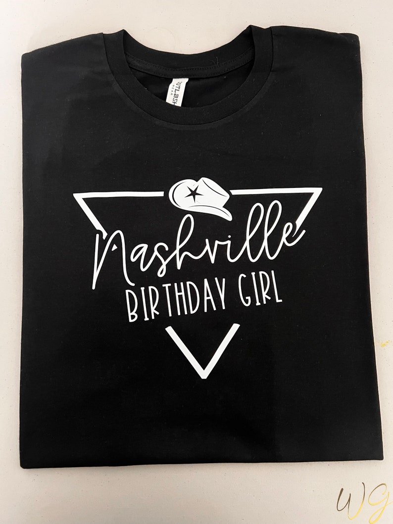 May include: Black t-shirt with a white graphic design of a triangle with a star and a cowboy hat inside. The text "Nashville Birthday Girl" is written inside the triangle.