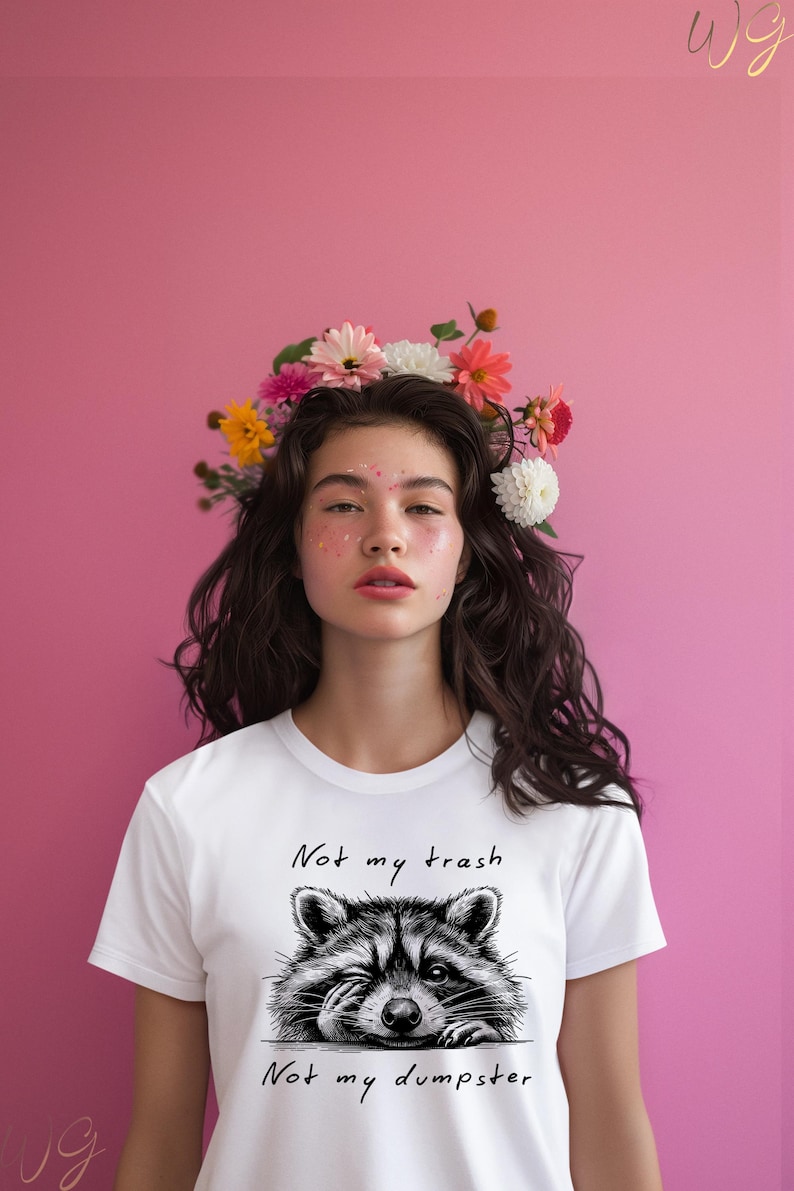 以下が含まれることがあります： 花冠をかぶった女性が、白のTシャツを着ています。Tシャツには、黒と白のラクーンドローイングと「Not my trash Not my dumpster」という文字がプリントされています。背景はピンクです。