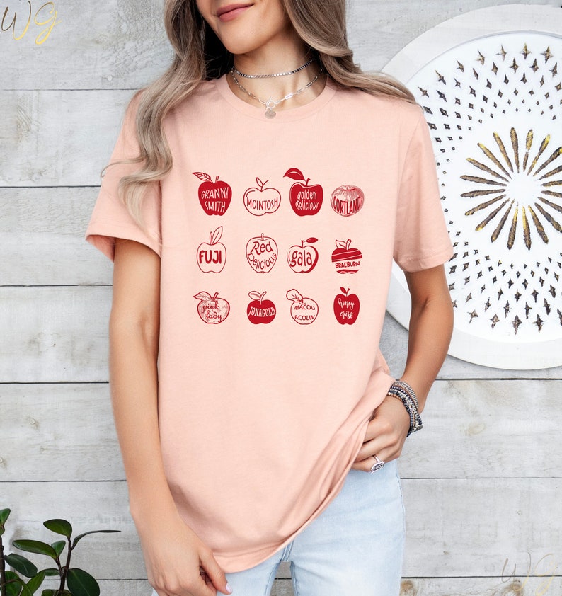 May include: A pink t-shirt with a graphic of red apples and their names, including Granny Smith, McIntosh, Golden Delicious, Cortland, Fuji, Red Delicious, Gala, Braeburn, Pink Lady, Honeycrisp, and  Macoun. The graphic also includes the text "Yummy Time".