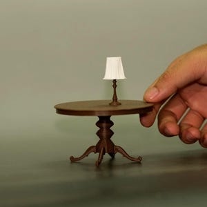 Puede incluir: Una mesa marrón en miniatura con una lámpara a juego. La mesa tiene una tapa redonda y patas ornamentadas. La lámpara tiene una pantalla blanca y una base marrón. La mesa tiene unos 25 cm de diámetro.