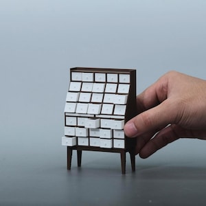 May include: A miniature brown wooden cabinet with numerous small white drawers. The cabinet has four legs and is being held by a hand. The drawers are arranged in a grid pattern, suggesting a storage or organizational function.