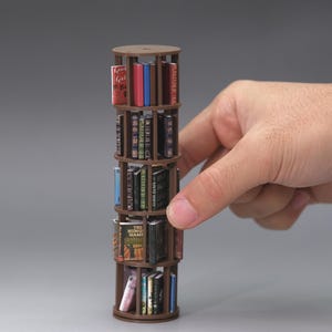 May include: A small, brown, rotating bookcase filled with miniature books. The bookcase has four levels and is held by a hand. The books are of various colours and titles, suggesting a collection of miniature literature.