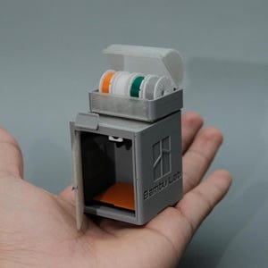May include: A gray 3D-printed storage container with an open door and a tray on top, holding several spools of filament in orange, white, green, and gray. The text "Bambu Lab" is visible on the side.
