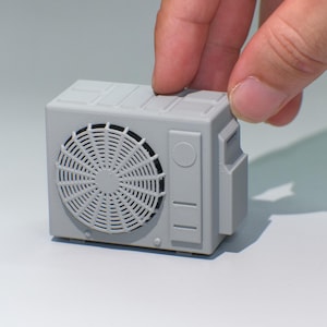 May include: A small, gray air conditioning unit model with a fan grill and rectangular design. The miniature unit is held by a hand, showcasing its compact size and detailed features. The model is a light gray color.