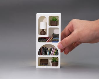 May include: A miniature white bookshelf with arched openings, containing tiny books, a lamp, and potted plants. The bookshelf is held by a hand, highlighting its small size. The lamp features a white shade and a brown base.