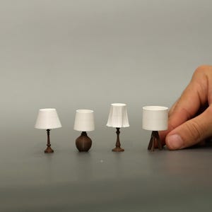 Puede incluir: Cuatro lámparas en miniatura con pantallas blancas y bases marrones. Las lámparas tienen diferentes diseños, incluyendo una forma clásica, una base redondeada, una pantalla plisada y una base de trípode. La lámpara más alta mide unos 5 cm de alto.