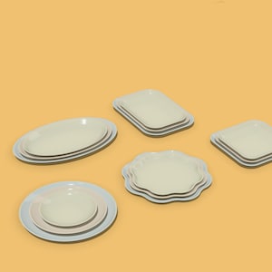 May include: A collection of ceramic dinnerware featuring various shapes including oval, rectangular, square, round, and scalloped-edge plates. Each set consists of stacked plates in shades of off-white and light blue, arranged on a solid yellow background.