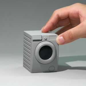 Puede incluir: Un pequeño modelo gris en miniatura de lavadora. La parte delantera presenta una puerta circular y detalles del panel de control. El lateral tiene detalles acanalados. El modelo está sostenido por una mano, lo que sugiere su pequeño tamaño. El diseño general es una representación minimalista de un electrodoméstico.